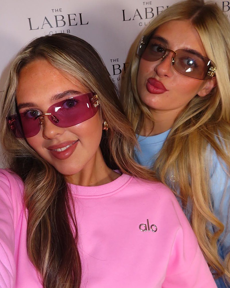 Two women wearing rimless designer sunglasses in front of The Label Club backdrop